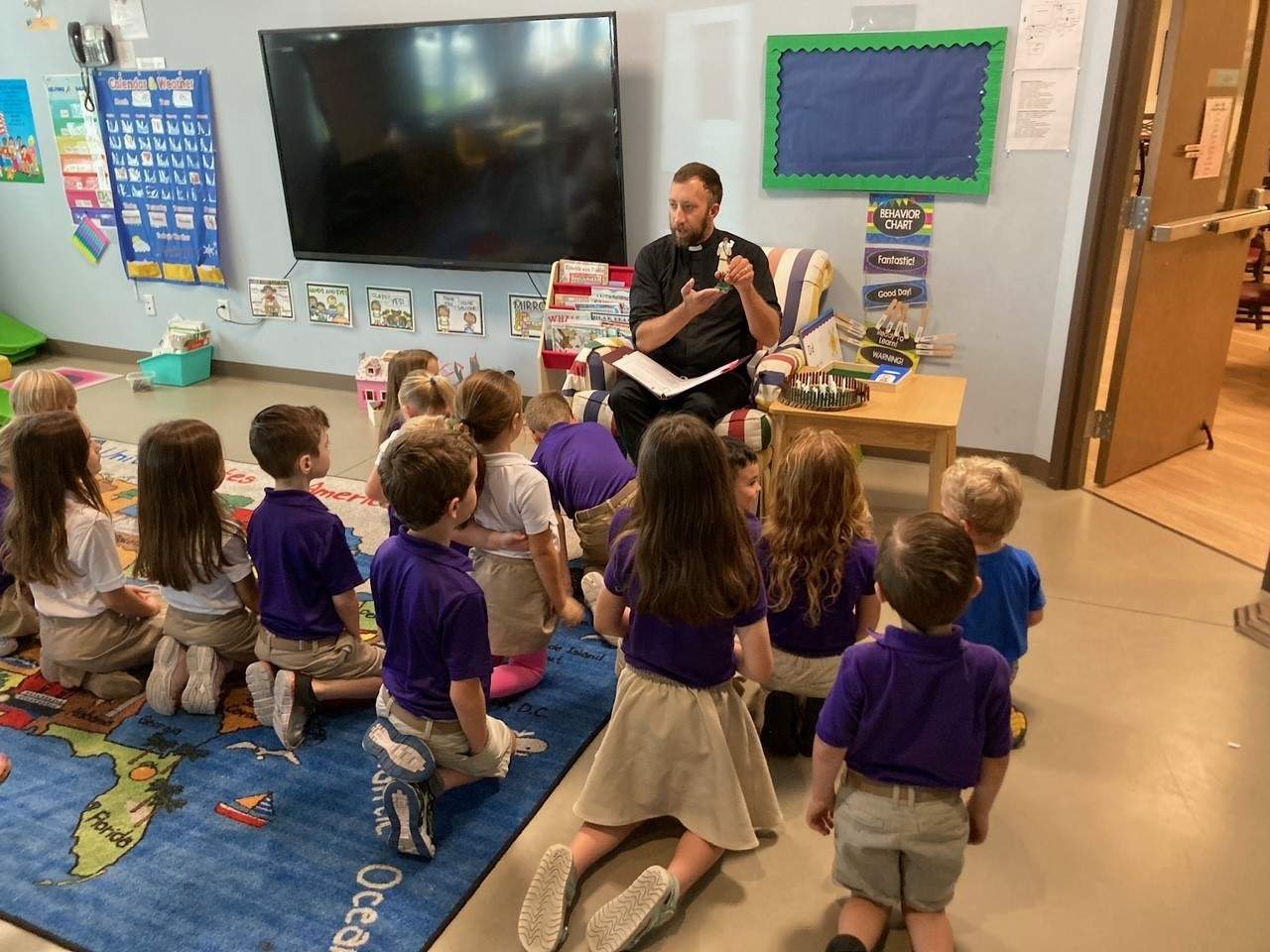 A teacher reading a book to a group of young students sitting on a classroom floor, all wearing purple uniforms.