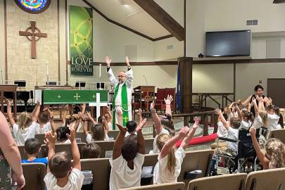 A pastor in green robes raises his arms, engaging with a group of children raising their hands inside a church hall.