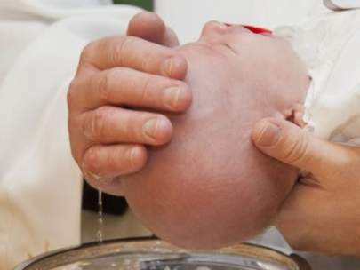 Close-up of adult hands holding a baby's head over a baptismal font, with water dripping from the baby's head.