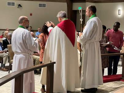 Clergy in white and green robes blessing a person in a church ceremony, with congregation members observing.