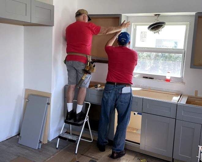 Two men installing upper cabinets in a kitchen, one standing on a ladder and the other assisting from the floor.
