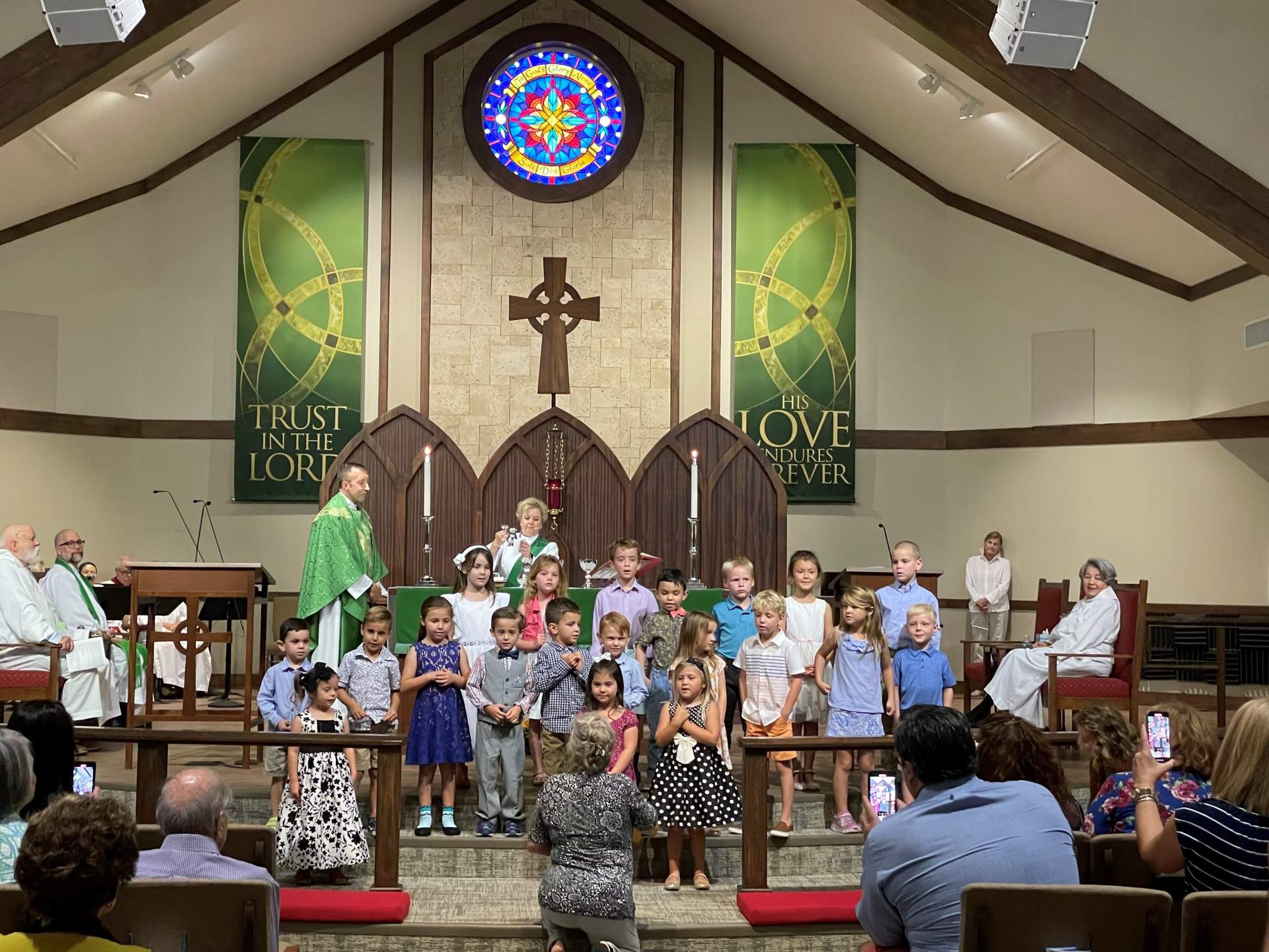 Children's choir standing in front of a church congregation, with adults seated and taking photos; stained glass window in the background.