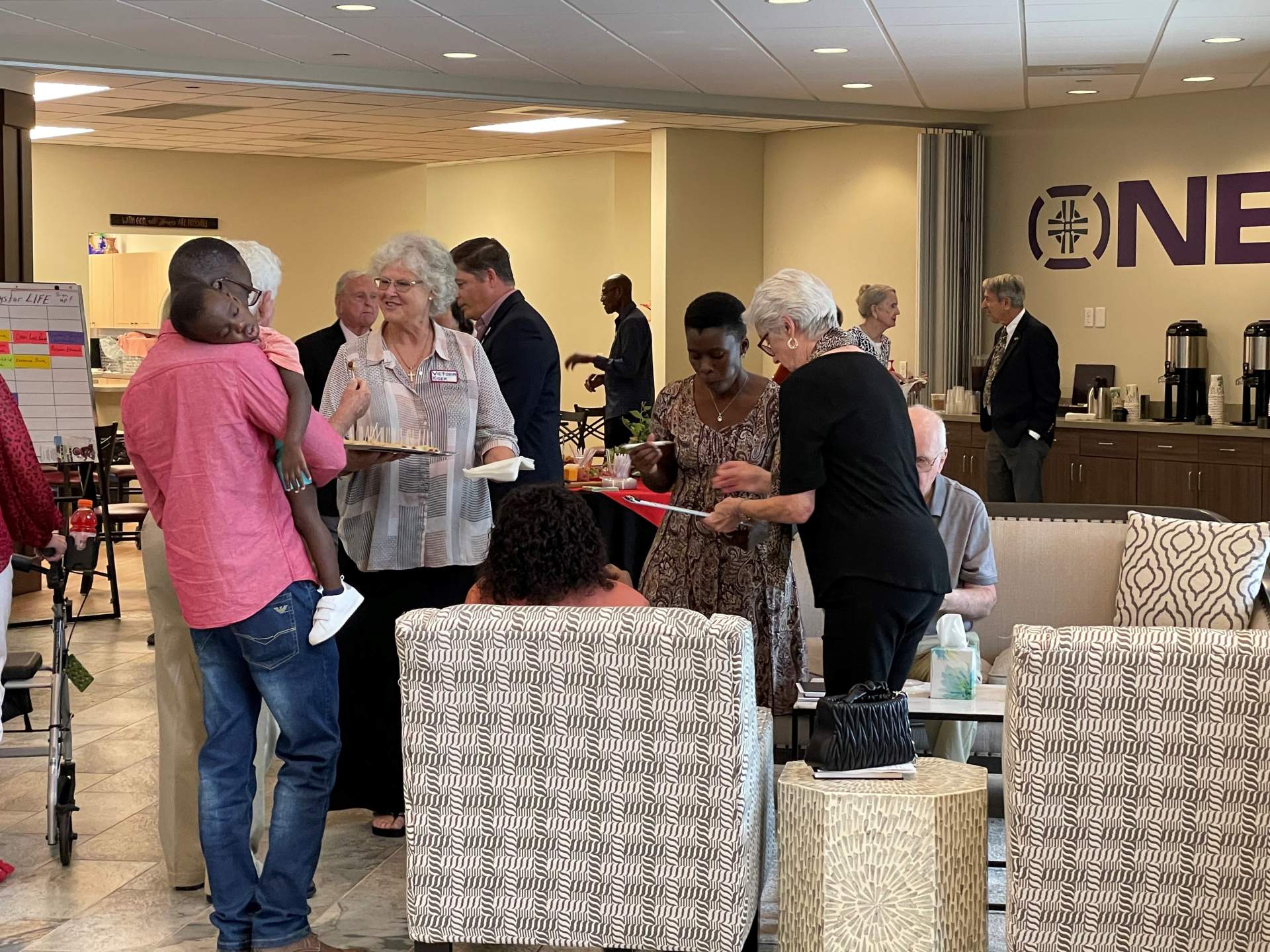 Diverse group of adults conversing and holding papers in a well-lit conference room with a coffee station in the background.